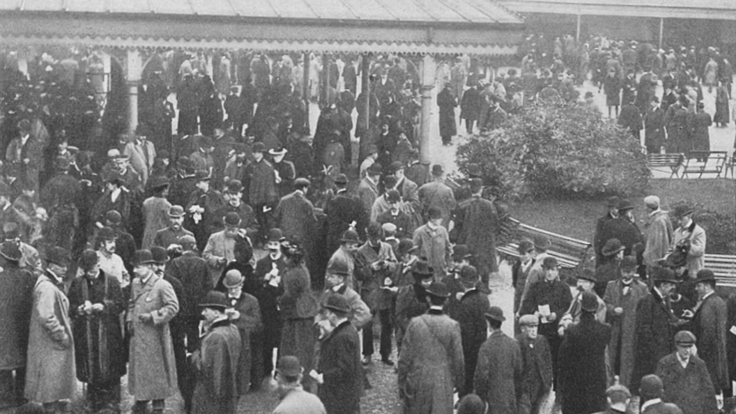Historic black-and-white photograph of the crowded racecourse enclosure with people dressed in formal Victorian-era clothing, including coats and bowler hats, gathered near a covered structure with ornate trim.