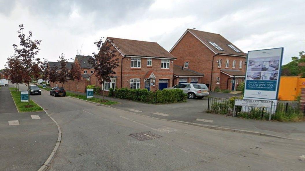 The entrance road to a housing development, with brick houses of different sizes on the right, some cars and a road sign. The road into the development is lined with red trees and and grass verges. 