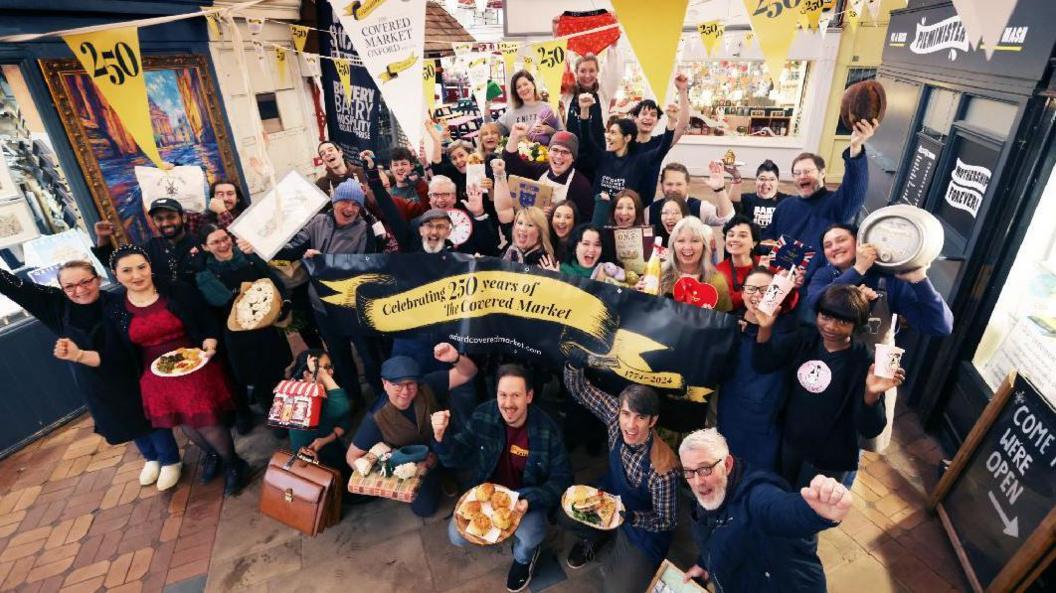 Traders of the Covered Market gather for a group photo, holding out their wares, such as cooked meals, baked items, milkshakes, flowers, and artwork. Banners and bunting proclaim the 25th-year anniversary.