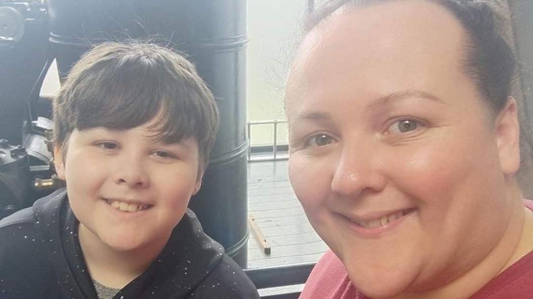 Claire and Dylan are looking at the camera and smiling in this selfie. Claire is wearing a red t-shirt with her hair tied back and Dylan, who has short dark brown hair, is wearing a black hoody with white speckles on it. 