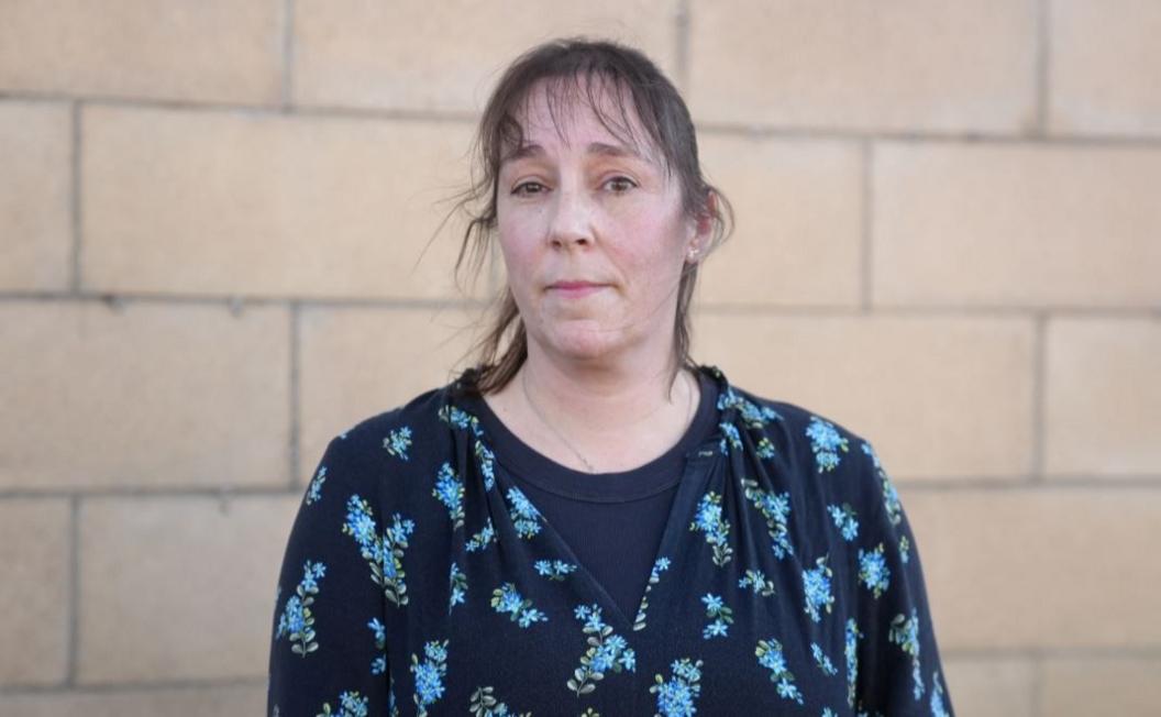 Julie Morgan looking at the camera. She has a fringe and dark hair and is wearing a navy top with blue flowers on, and is standing in front of a brick wall.
