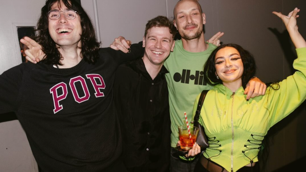 Four people posing for a photo, Charli XCX is wearing a lime green corset hoodie, the other people from left to right are wearing a black jumper with the word POP in red letters, a black shirt and another a green jumper