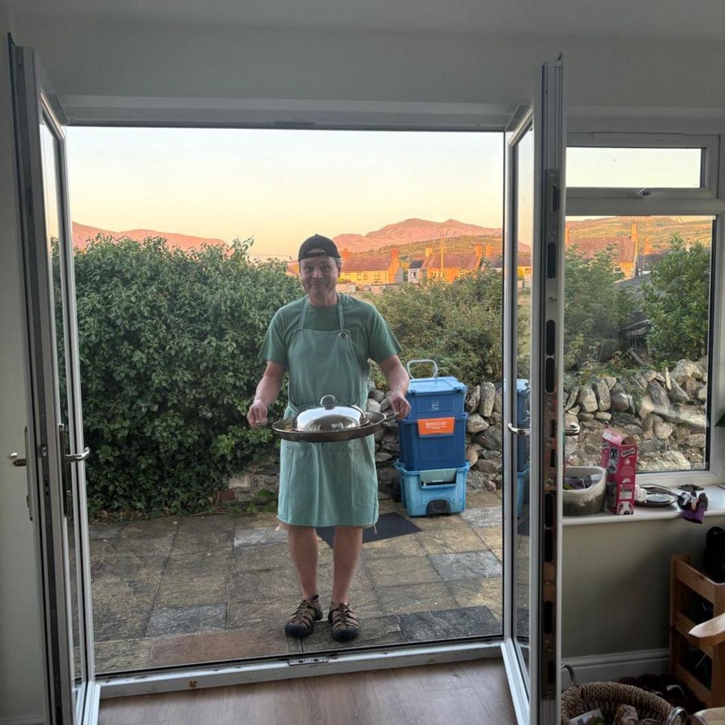 Dyn yn sefyll yn gafael ar bryd bwyd gyda mynyddoedd Eryri tu ôl iddo yn y pellter