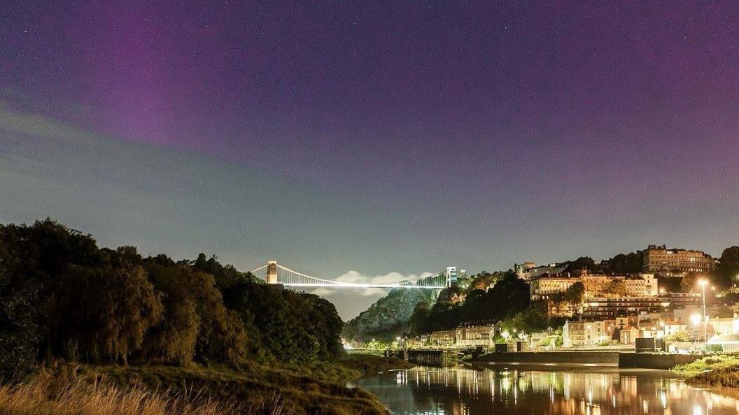 The northern lights are visible over the Clifton Suspension Bridge, with the lights of the bridge and Clifton reflected in the water of the River Avon below. The northern lights are a mixture of purple and green