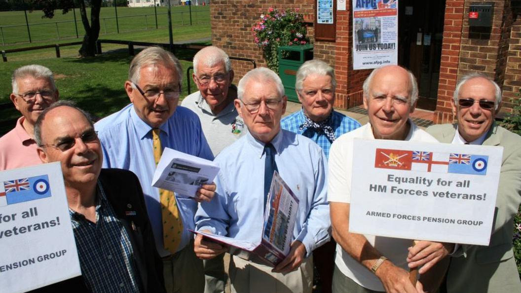 A photo from 2009. Eight men are stood in a group. Two of them hold posters that say Equality for all HM Forces Veterans. Peter Bottomley MP is third from left.