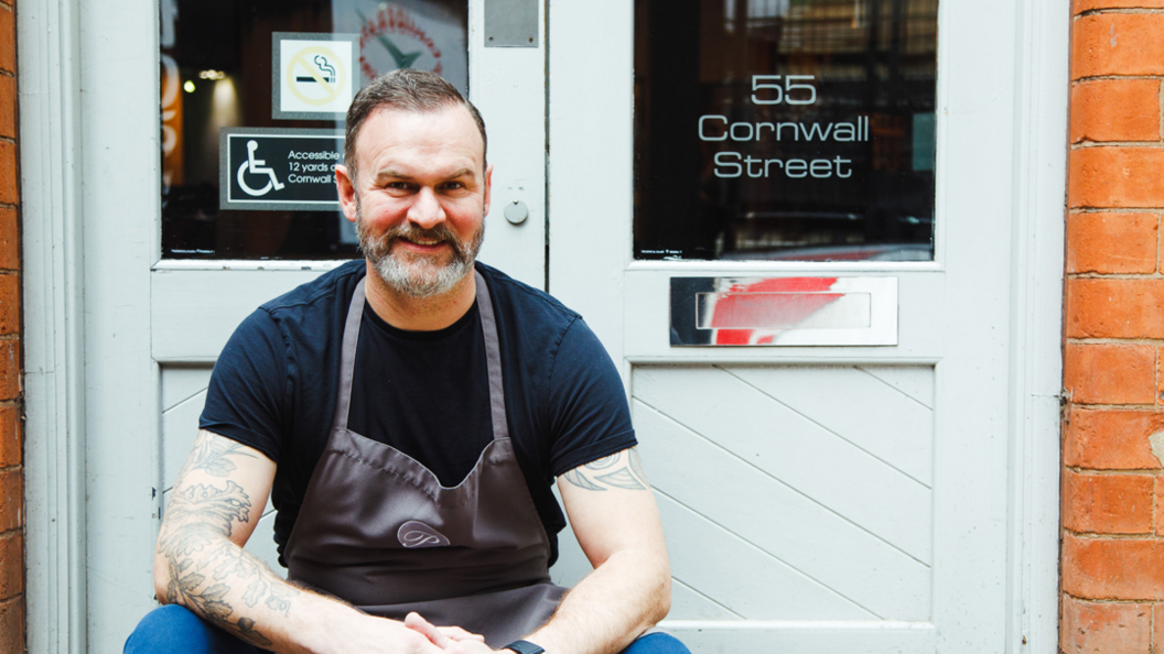 Glynn Purnell in a black t-shirt and apron, sat outside a white door at the front of his restaurant in Birmingham.