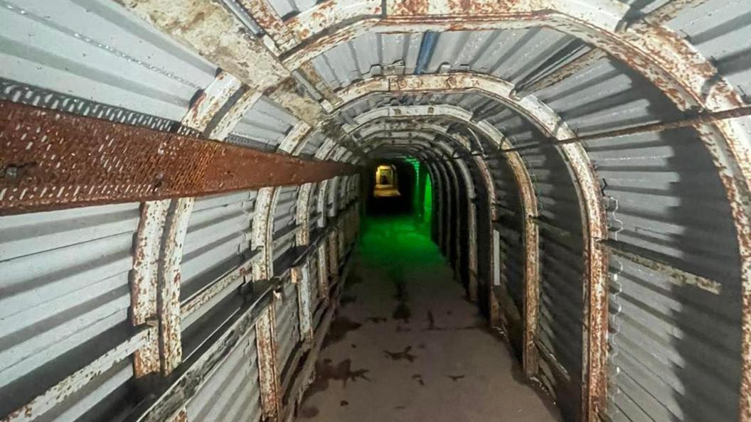 A long narrow tunnel lined with corrugated steel with a green light shining half way down