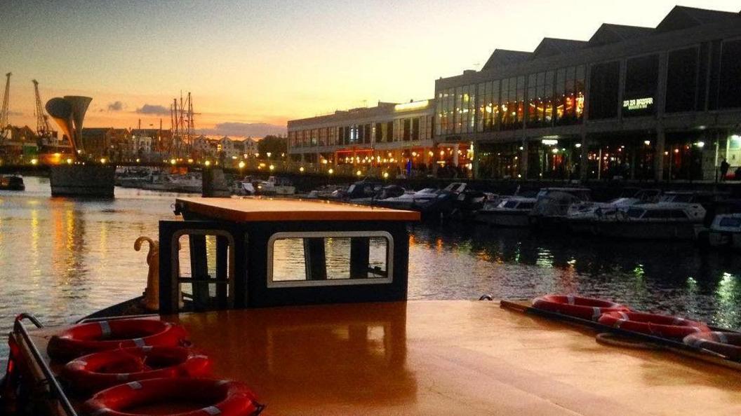 A Bristol Ferry Boat company vessel heads towards Pero's Bridge in Bristol city centre. The picture is taken at sunset and the lights of various bars and restaurants can be see reflected in the water of the floating harbour