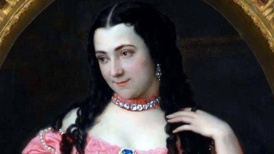 Portrait of a woman, dating from the mid 19th Century. She has a pale face, long black hair, parted in the middle and is wearing what appear to be diamond earrings and a pink choker with diamonds or glittering jewels. Her pink dress is off the shoulder and there is a blue jewel in the middle of the neckline. Her left hand is on her shoulder resting on her hair.