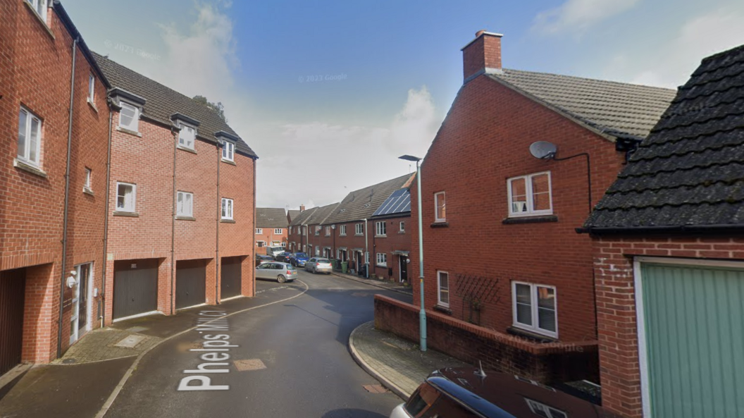 A streetview image of Phelps Mill Close. It is a relatively new looking street with a mix of taller houses over garages and terraced houses, all in red brick. 