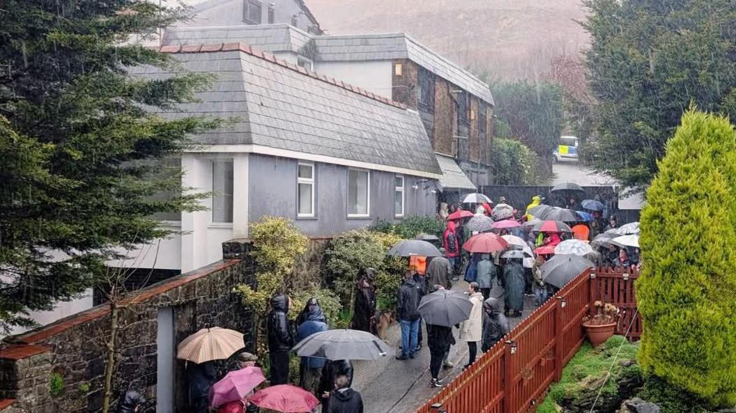 Picture of around 40 people gathered outside a home smiling towards a camera. Most can be seen stood in the rain wearing rain coats and carry red, black and grey umbrellas.