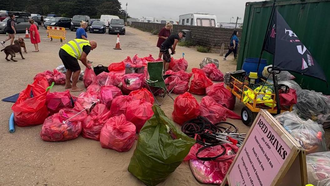 A sandy path covered in red bin bags which are full of rubbish., A couple of volunteers in high viz are visible, as well as some members of public and dogs in the background. In the background is also a caravan park.
