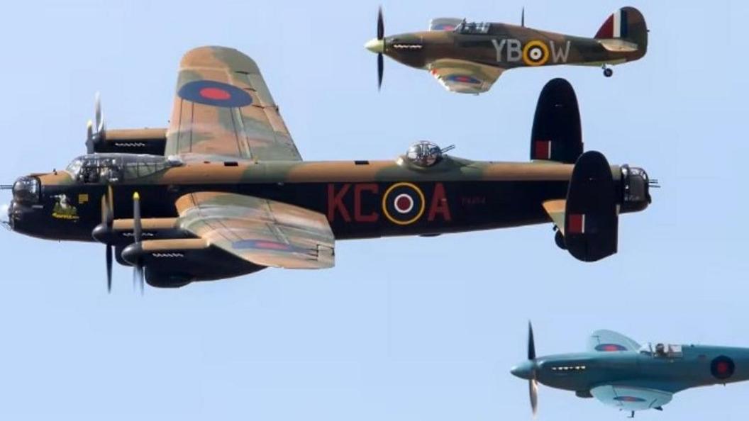 A Lancaster Bomber is flanked by Hurricane fighter plane and Spitfire. The Lancaster and Hurricane have brown and green camouflage markings while the Spitfire has marine blue.
