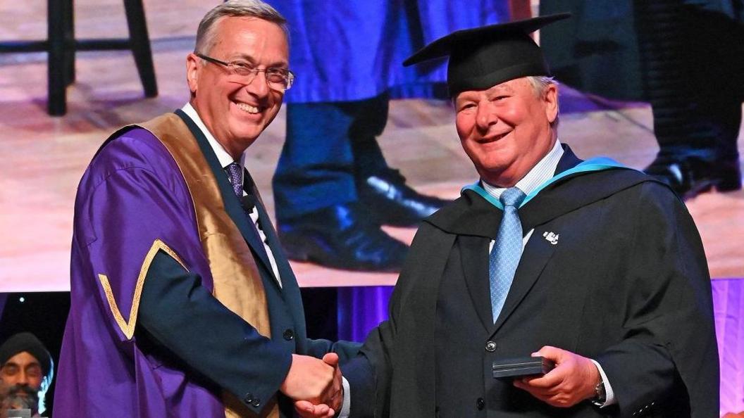 Vice-chancellor Stuart Croft presents the benefactor's medal to Tim Hartnoll in a ceremony at the university. Both men are wearing academic robes. They are smiling and shaking hands.