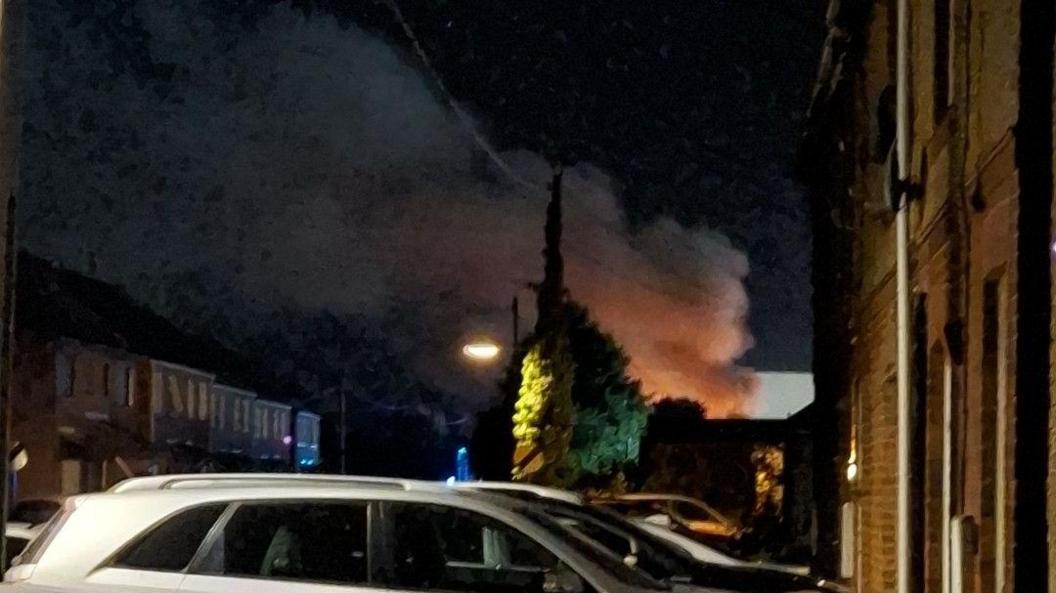 The picture of a housing area taken at night-time. There are cars and vans parked on both sides of the street. In the background, a plume of orange smoke is billowing into the sky.