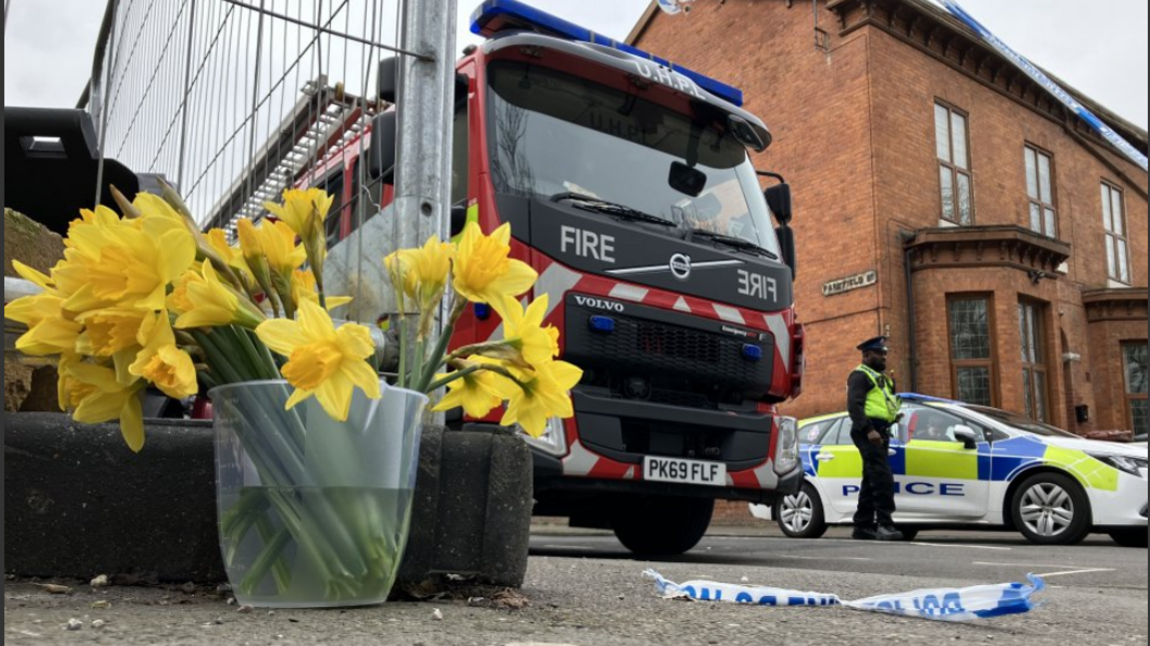 Daffodils in a bucket by the scene of the fire with a fire engine and a policeman also by the cordon