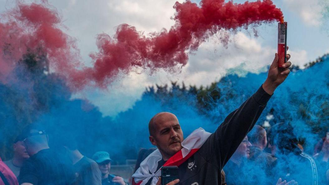 A man holds a red flare during a protest outside The Bell Hotel in Epping. He has a St George's flag draped over his shoulders.