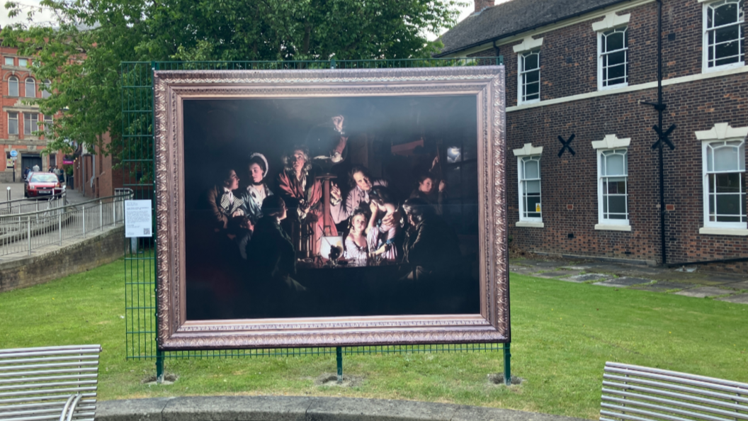A very large reproduction of a painting, set in the grounds of a park. The painting is largely dark, and features a number of people crowded together.