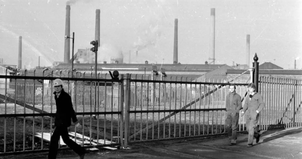 A black and white photo shows three men walking past metal gates. Behind them are various smoking chimneys of the Corby steelworks. 