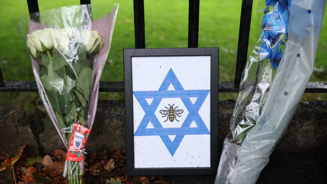 Floral tributes and the Star of David are displayed at the attack site. The tributes have been placed on the ground and are resting next to a black metal fence. Green grass can be seen behind the gate.