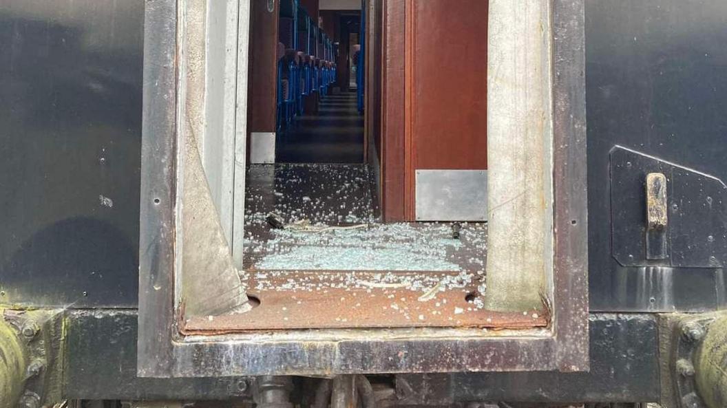 Damage to the side of a train carriage - a window is smashed and the smashed glass can be seen lying on the floor of the carriage