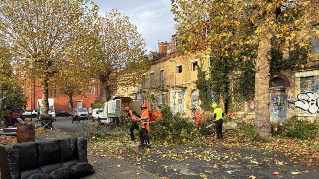 To the right of the image stands a row of derelict ochre-brick homes, with graffiti across the facade, boarded-up front doors and broken upper windows. Workmen in hi-vis jackets can be seen cutting back vegetation outside the terraced homes. In the foreground, to the left, lies a discarded black sofa.