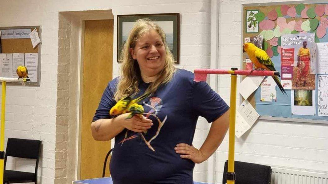 Eleen stands next to a yellow bird stand, she holds two yellow and green birds. Another yellow and green bird is on the perch next to her. Eleen has long blonde hair and wears a blue t-shirt.