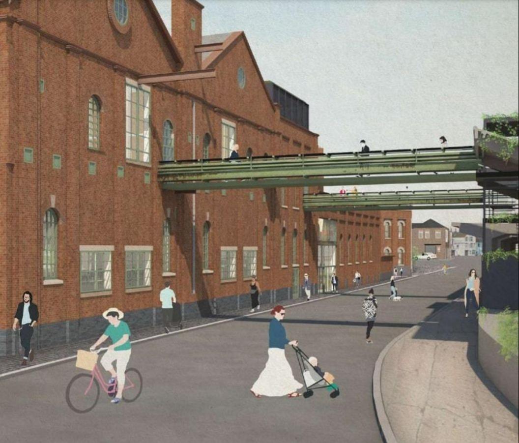 A large red-brick factory building from the late Victorian age, with glass windows of various shapes and sizes dotted along its length. Two green industrial conveyers, repurposed as bridges, cross high above a road, connecting the building to a structure on the other side. People walk and cycle beneath them.