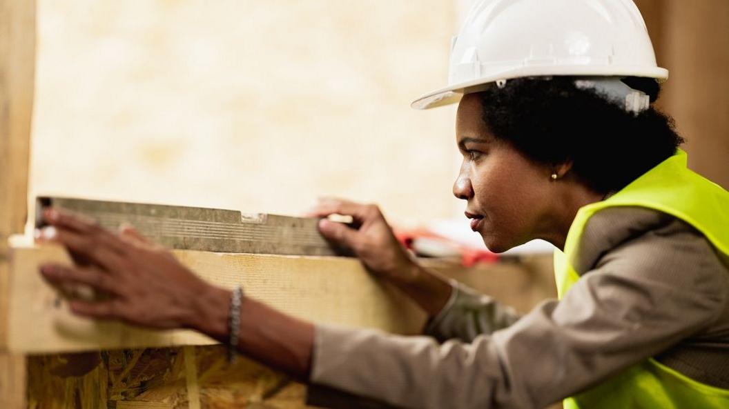 A woman with dark hair wearing a white hard hat, a grey top and green high-vis waistcoat, holds a piece of metal on top of the wooden frame of the window and stares at it