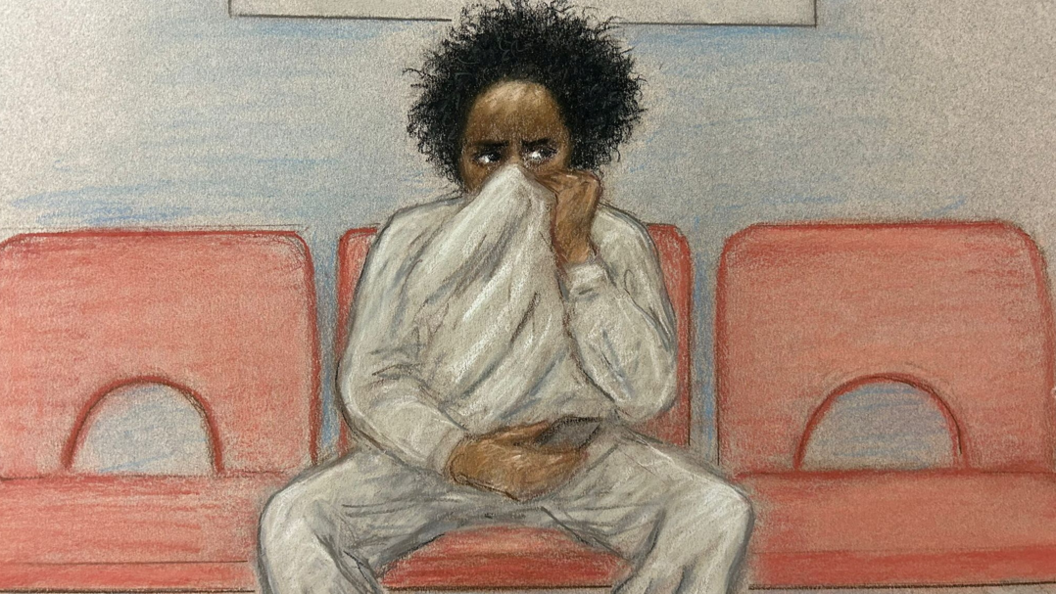 A court sketch of Axel Rudakubana sitting on a red chair. He has black, curly hair and is wearing a grey sweatshirt and joggers. He has his sweatshirt pulled up over his face, only his eyes are visible. 