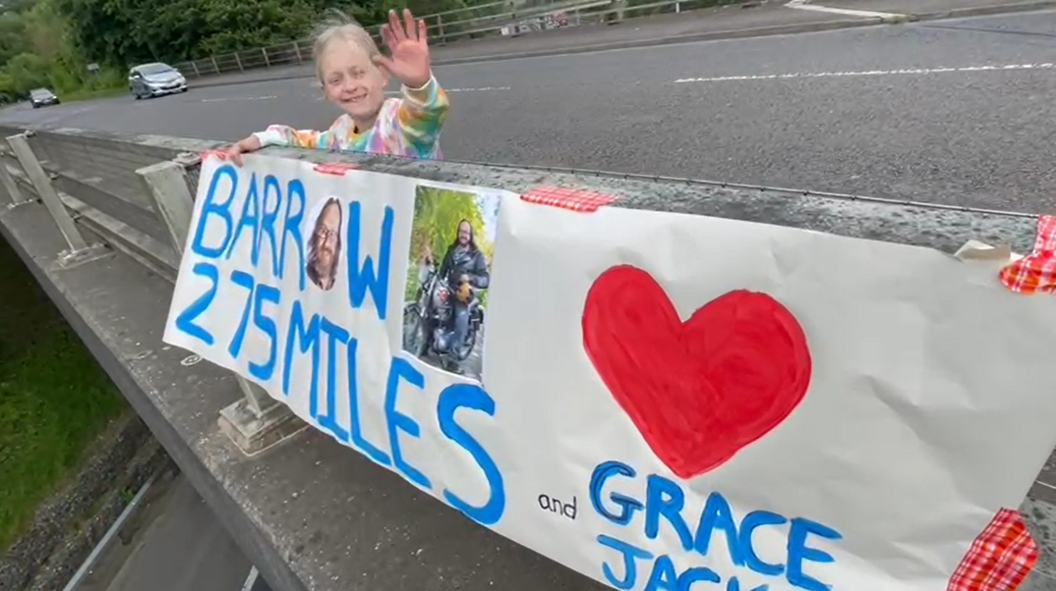 Girl waving to bikers with a sign that says '275 miles to Barrow'