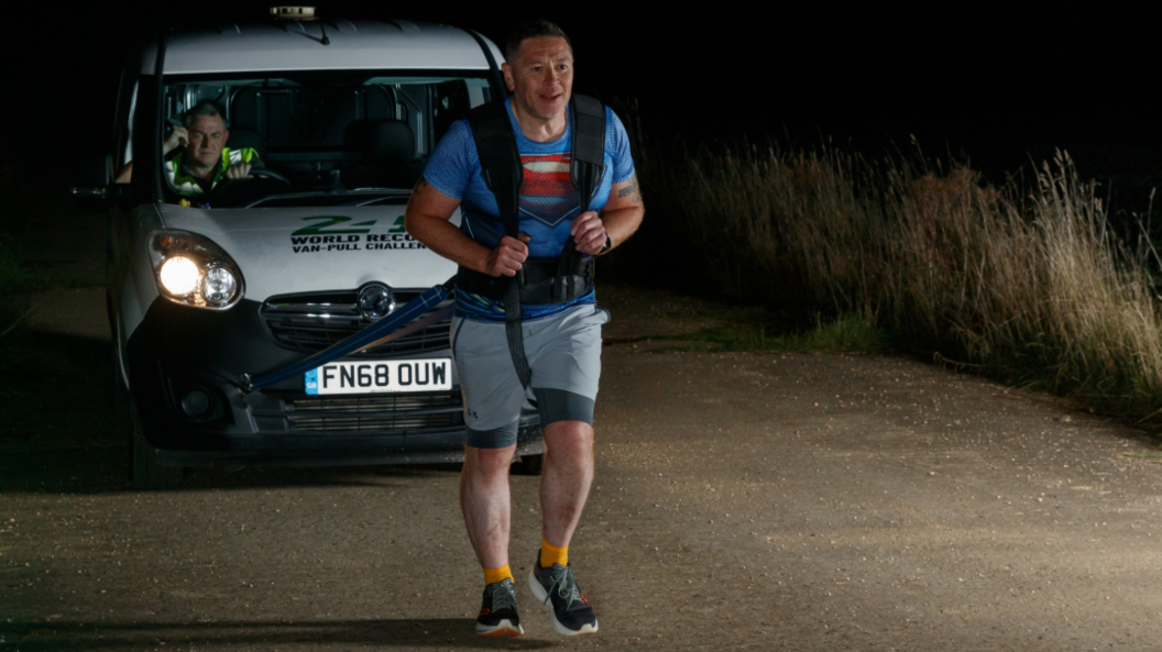 A photo taken at night of last year's record attempt with Mike, wearing a Superman Tee shirt, strapped to a white van and walking in front of it. He is being lit up by the van's headlights.