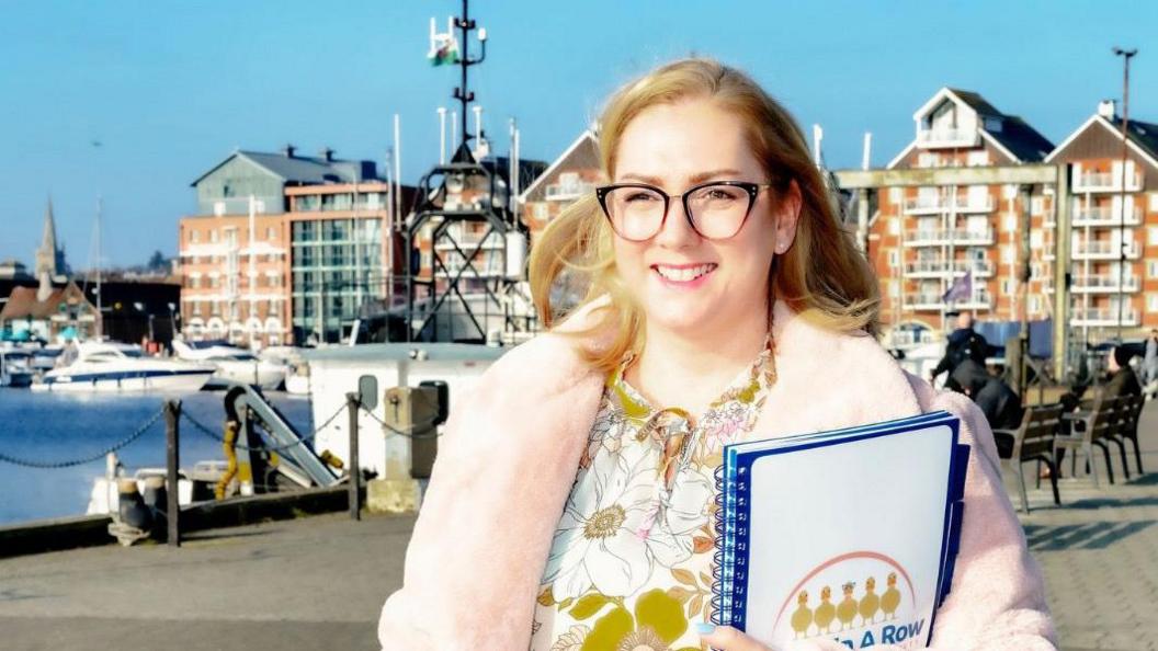 Clare Lucas walks along a promenade smiling. She holds a paper pad in one arm. She wears glasses, a pink fluffy coat with a green and white floral top underneath and she has long blonde hair.