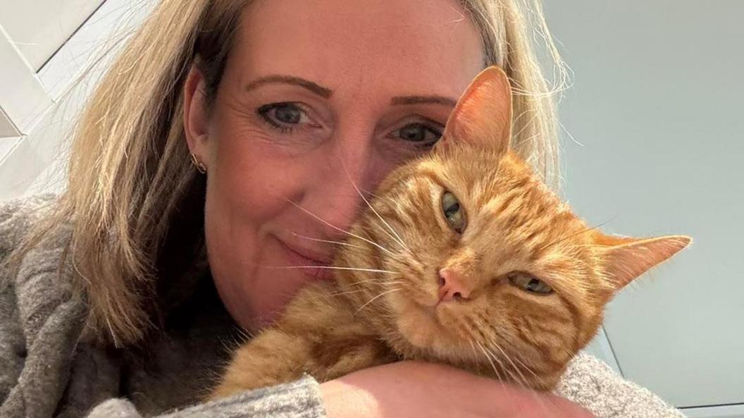 A woman with blonde hair wearing a beige jumper holds a ginger cat