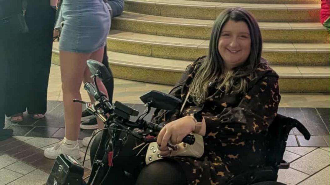 Sophie Shuttleworth smiles at the camera, she is sat in a wheelchair and people are visible behind her. 