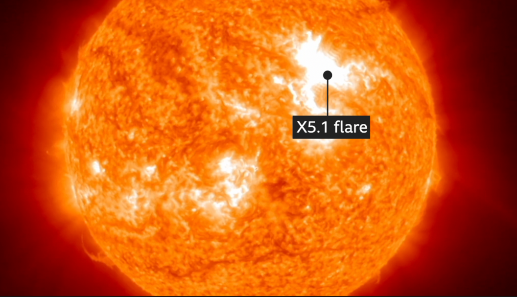 A close-up image of the Sun’s surface, showing a bright orange colour with white sunspots and an active solar flare region featuring a white burst.