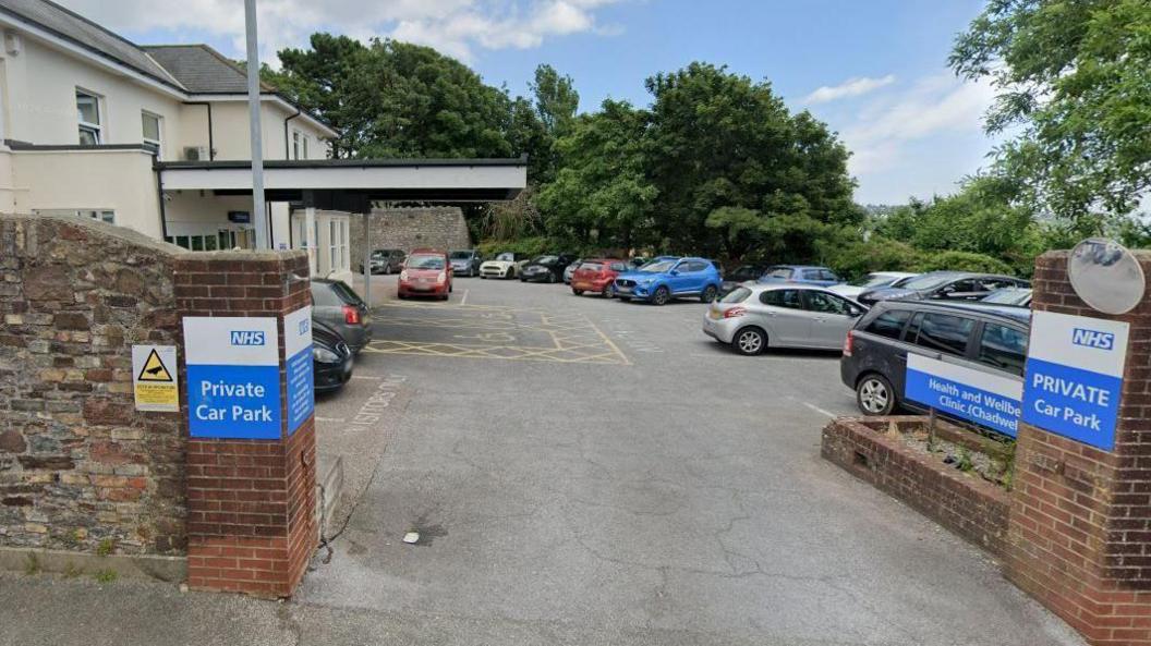 The Chadwell Centre in Paignton. The car park outside the white and cream-coloured building is filled with vehicles. A brick wall is on the perimeter of the car park. There are blue and white NHS signs saying the car park is private. Trees are dotted around the opposite side of the car park.