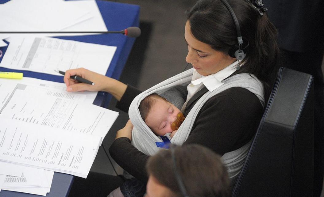 In a photo from 2010, European deputy Licia Ronzulli sits in parliament and signs papers while she holds her baby who is asleep in a sling, sucking a pacifier