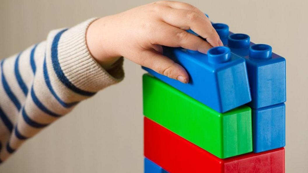 A young child's arm reaches up to a pile of red, blue and green bricks