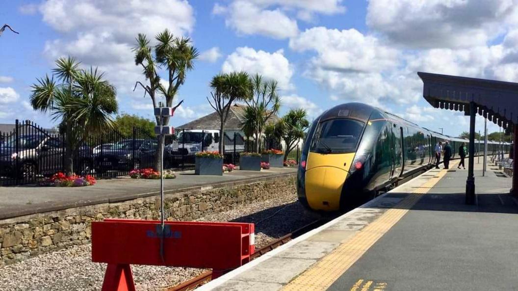 The trees at their previous home beside Newquay train station. They are on the far side platform of a railway station. The trees have flowers planted at their base of varying colours. There is a train pulled into the station, which has many carriages going off in to the distance. People are boarding the train. 