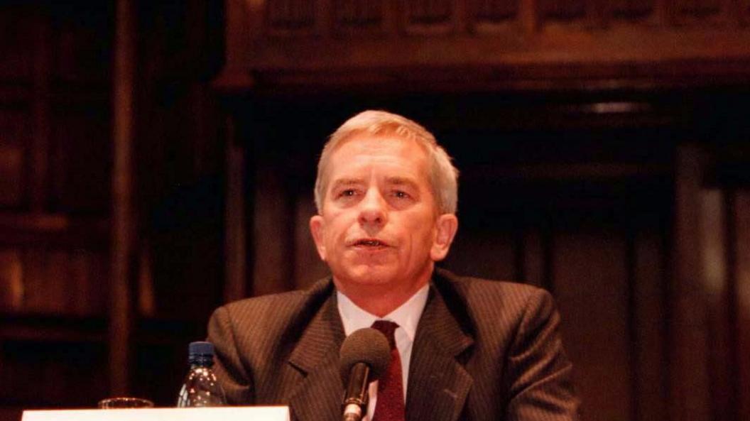 Lord Saville speaking into a mic at a hearing.  He is wearing a striped blazer, white shirt and a red tie.