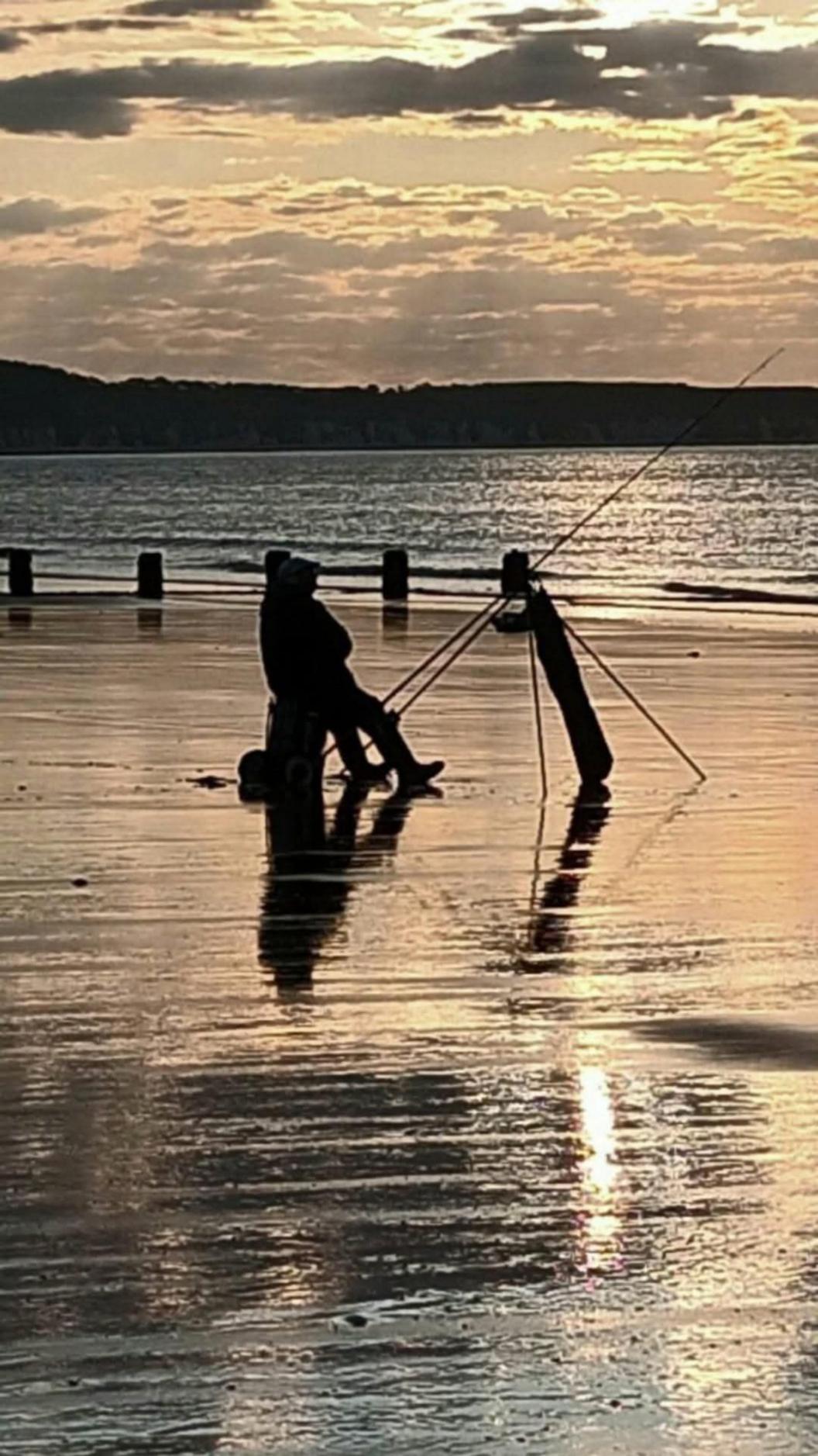 A photograph of a solitary fisherman on the beach. He is silhouetted. The majority of the image is sand, and then the sea can be seen - and some cliffs, as well as a few clouds. The photo has been taken at sunrise.  