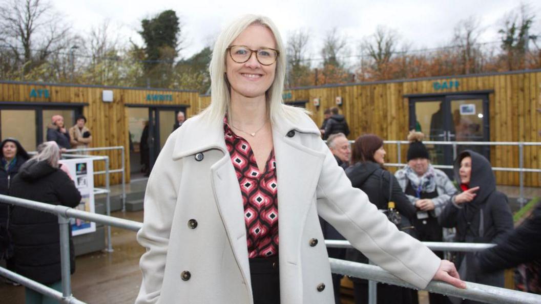 Emma Soanes smiling while standing outdoors at an event - there are platforms with wood panelling behind her and she is leaning on a railing, wearing a white coat and a red, white and black top.