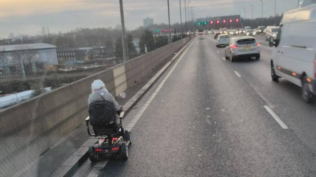 A busy raised dual carriageway, with a woman driving on the left hand side in a mobility scooter. The picture is taken from the window of the vehicle behind. Cars can be seen driving away in the lane to the woman's right, and in the direction towards her on lanes to the left. The mobility scooter is black. The woman has her back to the camera and is in a beige coat and cream hat.