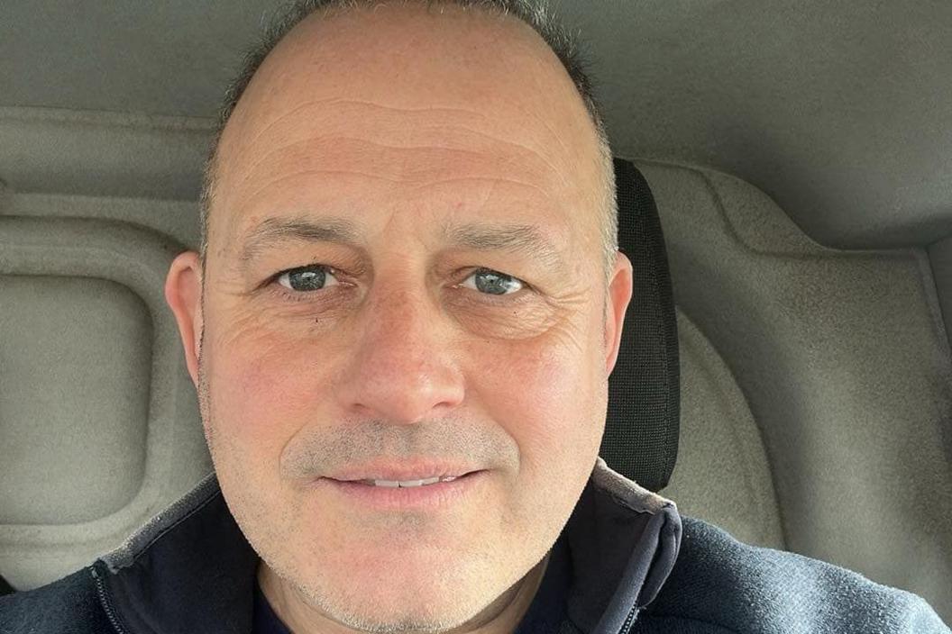 Man smiling in selfie style. He is balding and is sat in the car. He has blue/green eyes and is wearing a blue jumper. It is a head and shoulders shot.