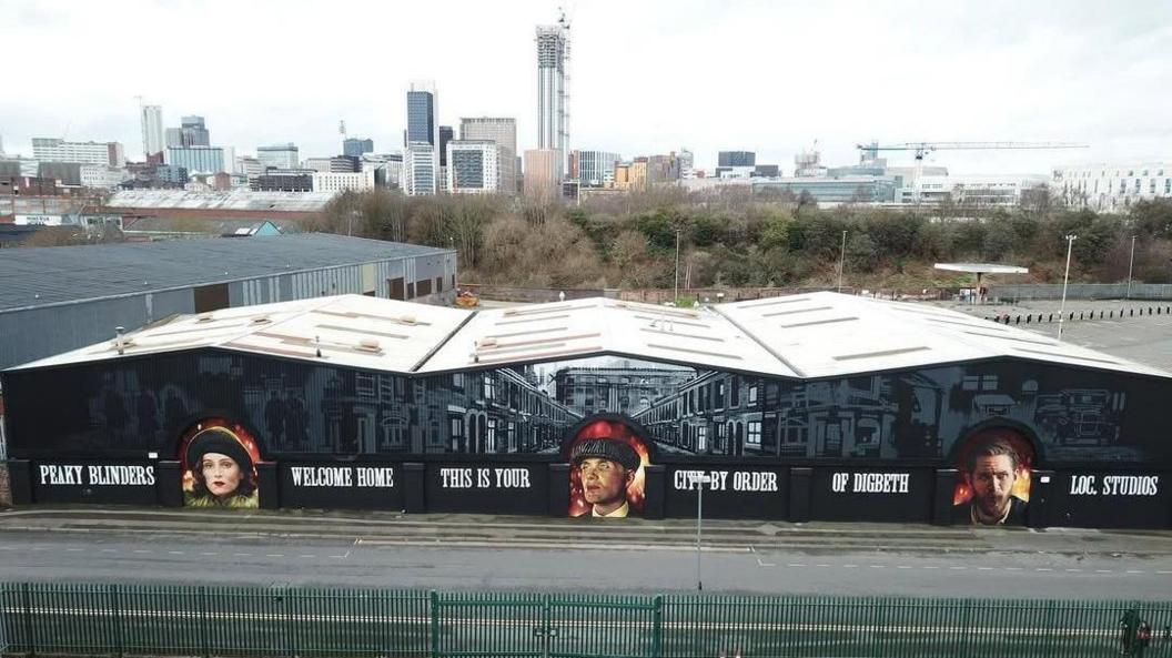 The outside of the Digbeth factory shows the three murals painted on the walls. The factory has a white roof and the words 'Peaky Blinders, welcome home, this is your city by order of Digbeth Loc Studios' painted on the walls.