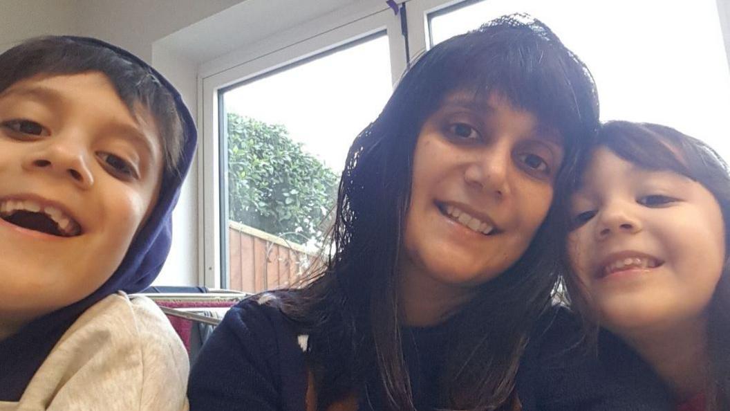 Sapna Boden with her two young children. She has long dark hair with a fringe, dark eyes and is wearing a blue jumper. Her head is titled towards her daughter on the right, and her son is on the left. They are all smiling at the camera and sitting in front of a bright window. 