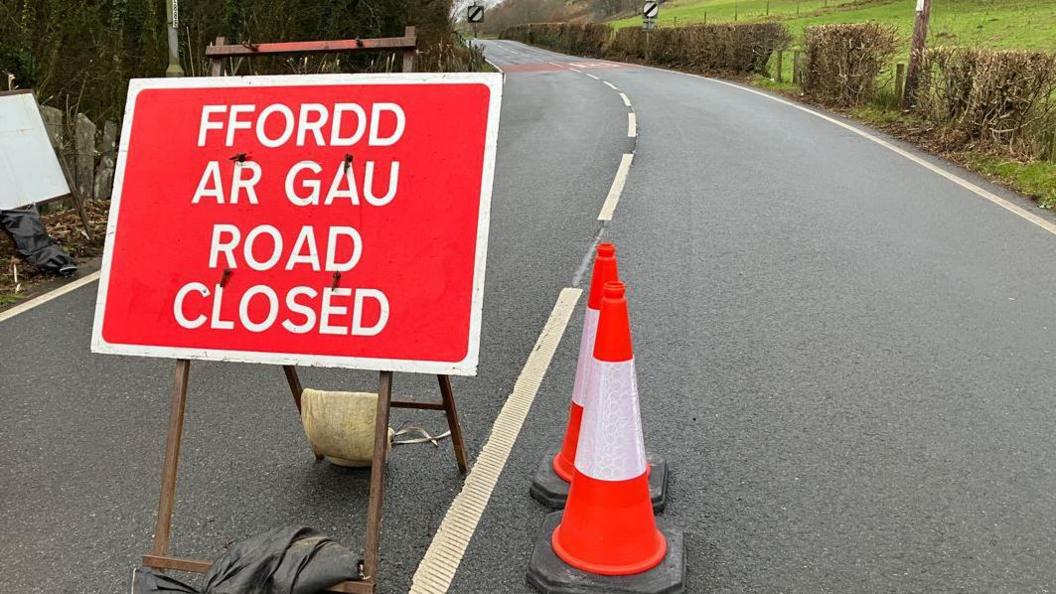 A red sign on a road stating it is closed. To the right of the sign are two cones. The road is in a rural setting