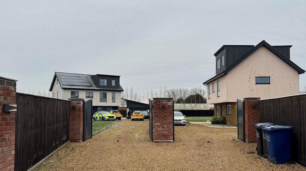 Two three-storey houses are set in a large shared gravel driveway. There are gates at the beginning of the drive and more than three police cars can be seen parked in front of the houses