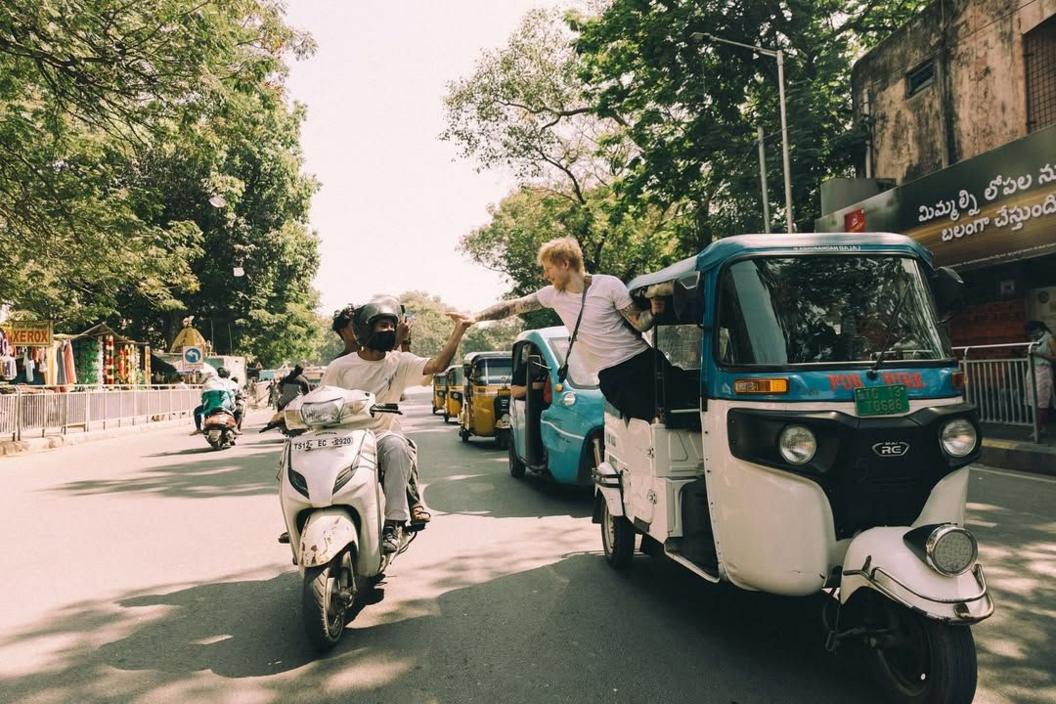 Ed Sheeran leans out of a Tuk Tuk and high fives two men riding alongside him on a scooter